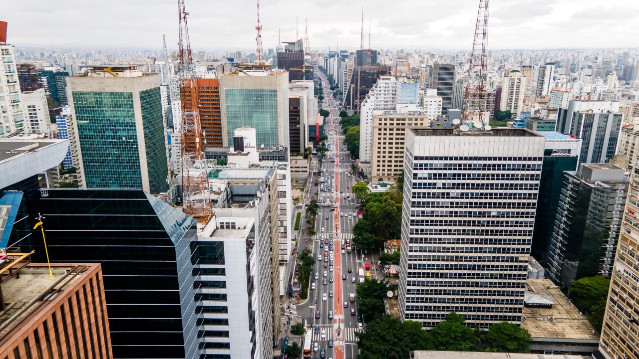 Foto de cidade - Entre muitos prédios e torres de comunicação, a avenida Paulista com as pistas cheias de carros  nos dois sentidos corta toda a imagem que é tirada do alto e em longa perspectiva, tendo uma grande área urbana na imagem